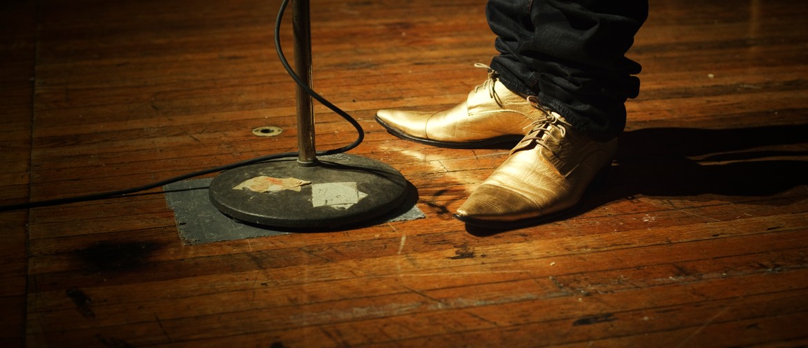 A person, only visible from the mid-calf down, wearing dark pants and gold lace-up pointy shoes, stands on a scuffed, wooden stage before a microphone stand; the microphone cord is wrapped around the stand and disappears off to the left of the frame. Not 
