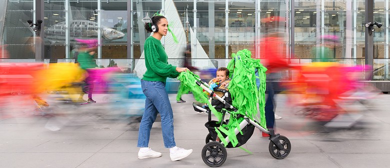 A Pram People photo. An adult in a green jumper and headphones pushes a small child in a pram decorated with green streamers. They are surrounded by colourful motion blurs. The adult looks out of frame, while the child looks at the camera. Photographer: S