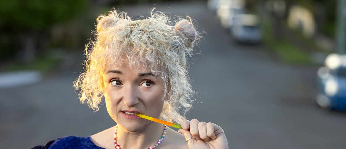 A girl with curly blonde hair in two buns bites into a rainbow lolly snake and looks to the side of frame. She wears a giant blueberry costume and a rainbow bead necklace, and stands in the middle of an empty road.
