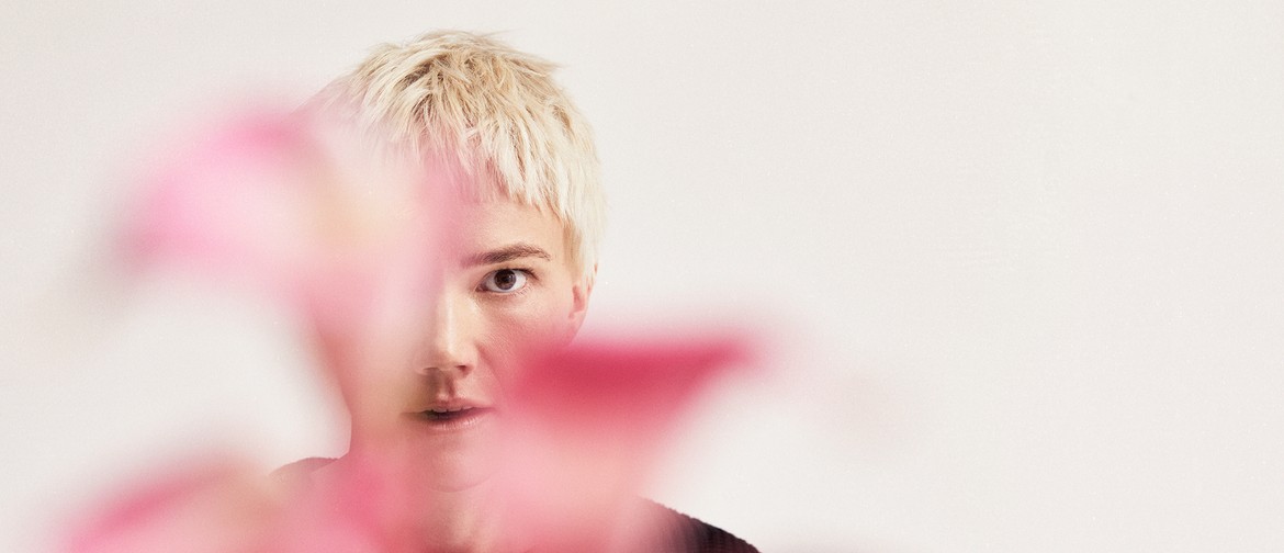 An image of a female-presenting performer with short, platinum blonde hair and an intense, mischievous expression stares at the camera - her face is partially obscured by vibrant pink out of focus flowers.