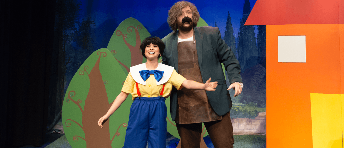 An actor dressed as Pinocchio in a black bowl cut wig, yellow t shirt, white collar, blue bow, and blue pants with red suspenders smiles widely and gesturing outwards with their arms. Another actor dressed as Gepetto in brown pants, a brown apron, and a g