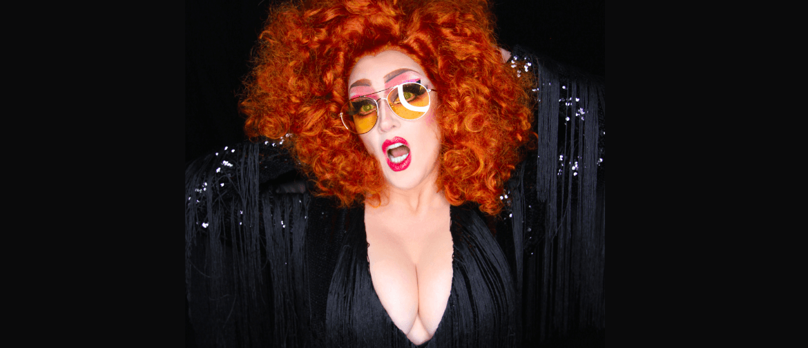 Drag performer with sunglasses on and red hair has mouth open to camera with cleavage showing. 