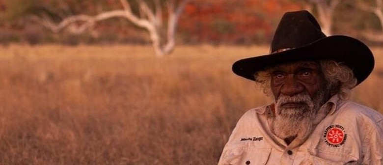 Aboriginal elder Nyarri Nyarri Morgan wears a khaki coloured ranger shirt and a black stockmans hat and sits out on country at dusk, with the trees and grass beginning to darken behind him.
