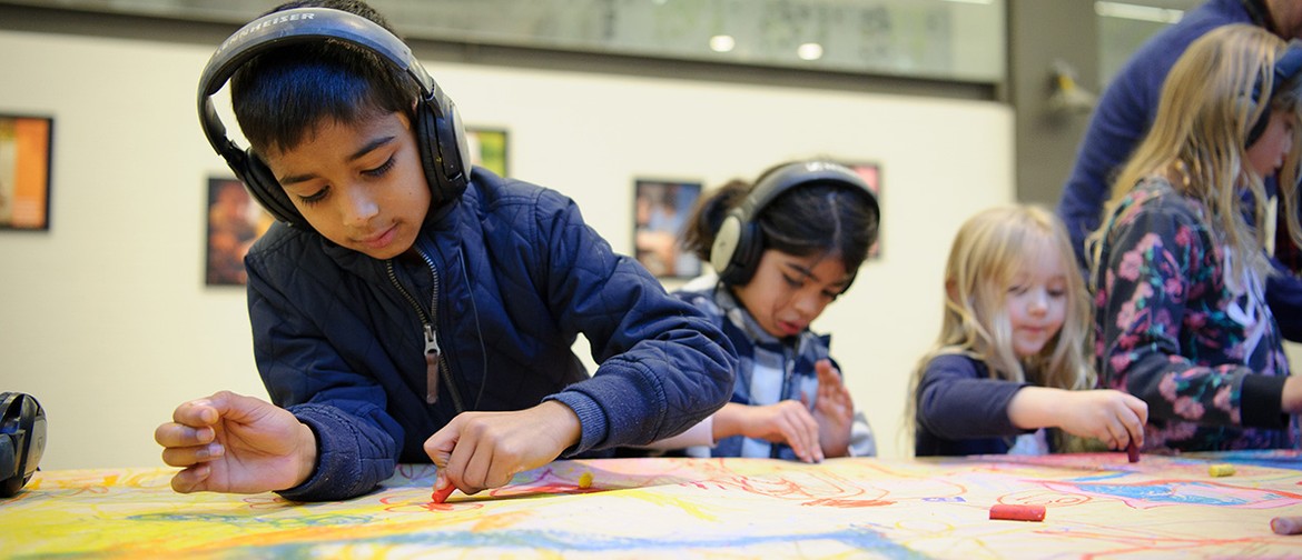 A Sound of Drawing production photo. A child wearing a jacket and headphones, draws intently on a table covered in paper. Other children are visible behind them, also drawing. Photo: Sarah Walker