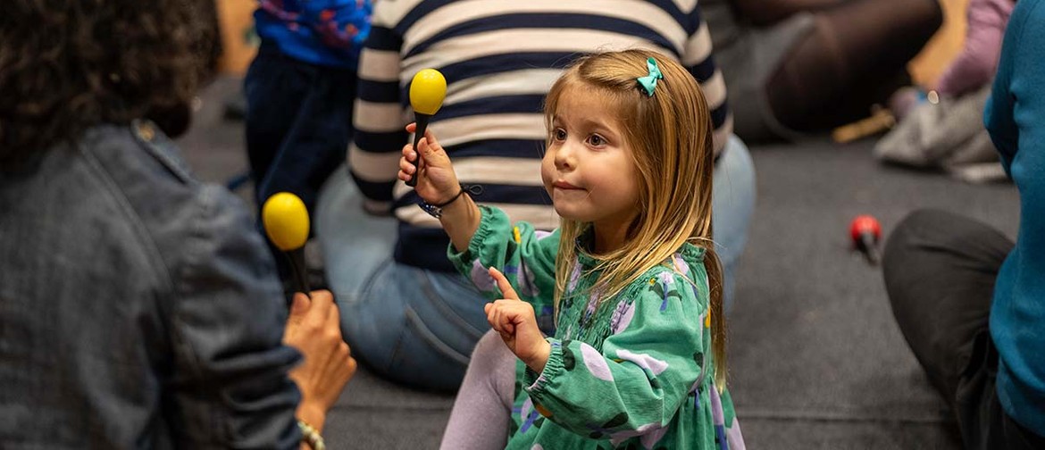 Jams for Juniors: Grieg's Piano Concerto A small girl, wearing a green dress with purple spots, holds a maraca in one hand and points with the other