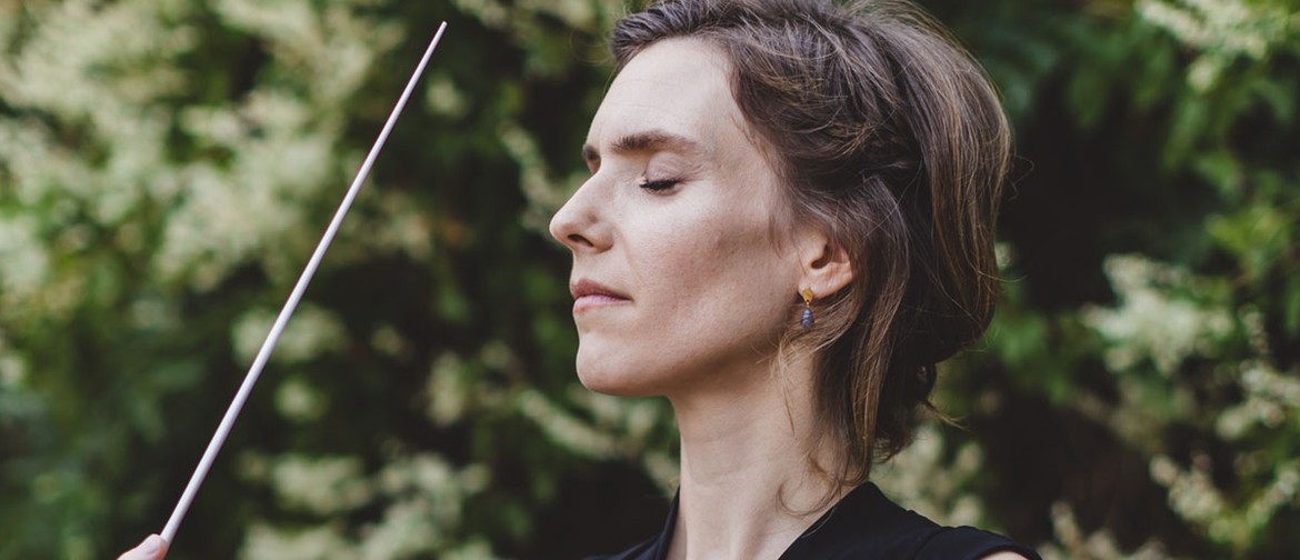 Franck's Symphony Conductor Chloé van Soeterstède stands in front of a rest backgrdop, eye closed, baton pointed skyward with her eyes closed looking serene