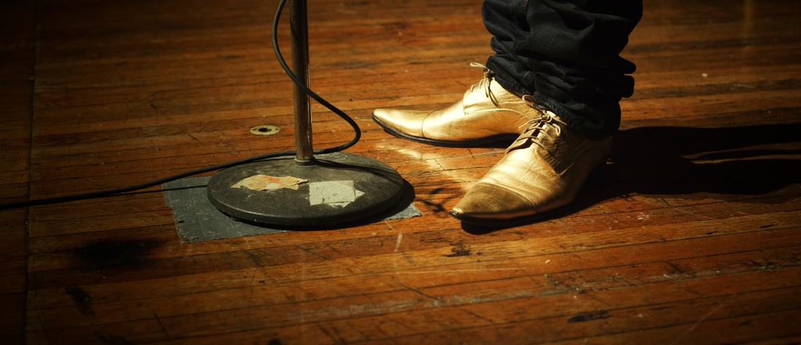 The Moth StorySLAM: DIRT A person, only visible from the mid-calf down, wearing dark pants and gold lace-up pointy shoes, stands on a scuffed, wooden stage before a microphone stand; the microphone cord is wrapped around the stand and disappears off to the left of the frame.
